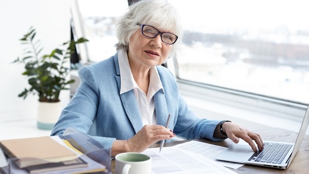 Attractive serious female chief executive officer of mature age sitting at her office with laptop, keyboarding and signing papers on desk, having confident look. People, aging, job and career concept